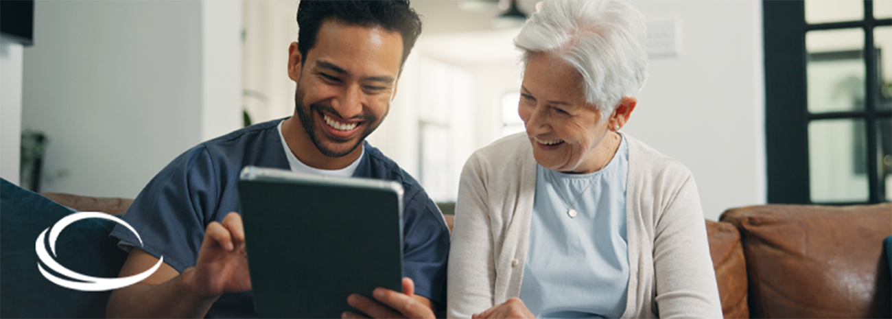 Healthcare professional sitting next to older woman on sofa and showing her some information on a tablet